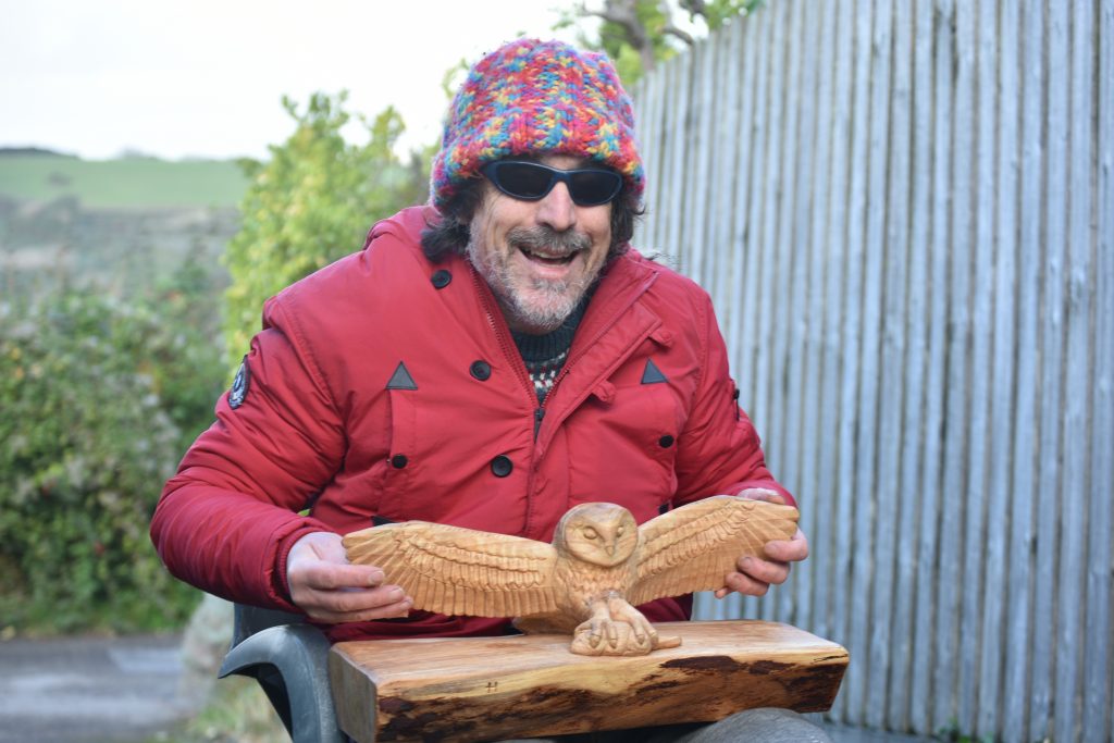 Bryan sits outside on a chair with barn owl carving on lap. He touches the wood carving, inspecting the shape. He wears his usual multi coloured woolly hat.